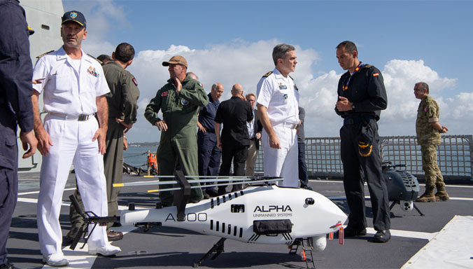 Thirteen uniformed personnel from different units converse during a NATO maritime exercise. A white unmanned helicopter drone is on the foreground, with the words 