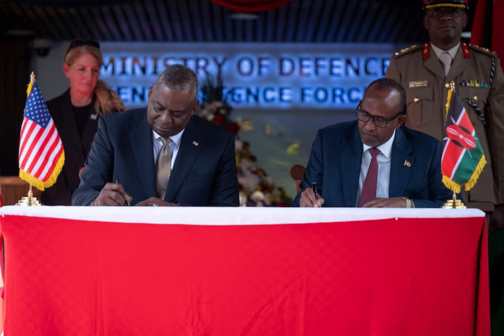 Secretary of Defense Lloyd J. Austin III (left) and Kenyan Defense Minister Aden Bare Duale (right) sit beside each other while signing the papers of the five-year framework for defense cooperation. They are flanked by one military personnel each on the background. A US flag is placed on Austin's side, while a Kenyan flag is on Duale's.