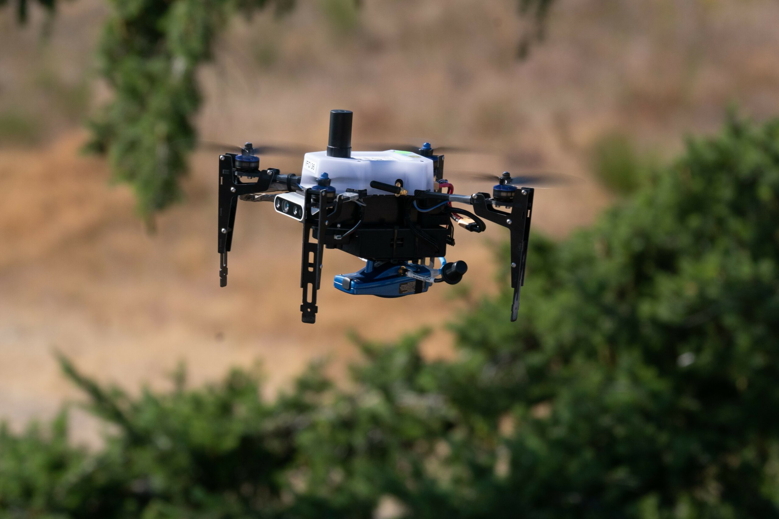 A small hovering drone is front and center in the picture. In the blurred out background, some greenery are seen in an otherwise rocky/sandy area.