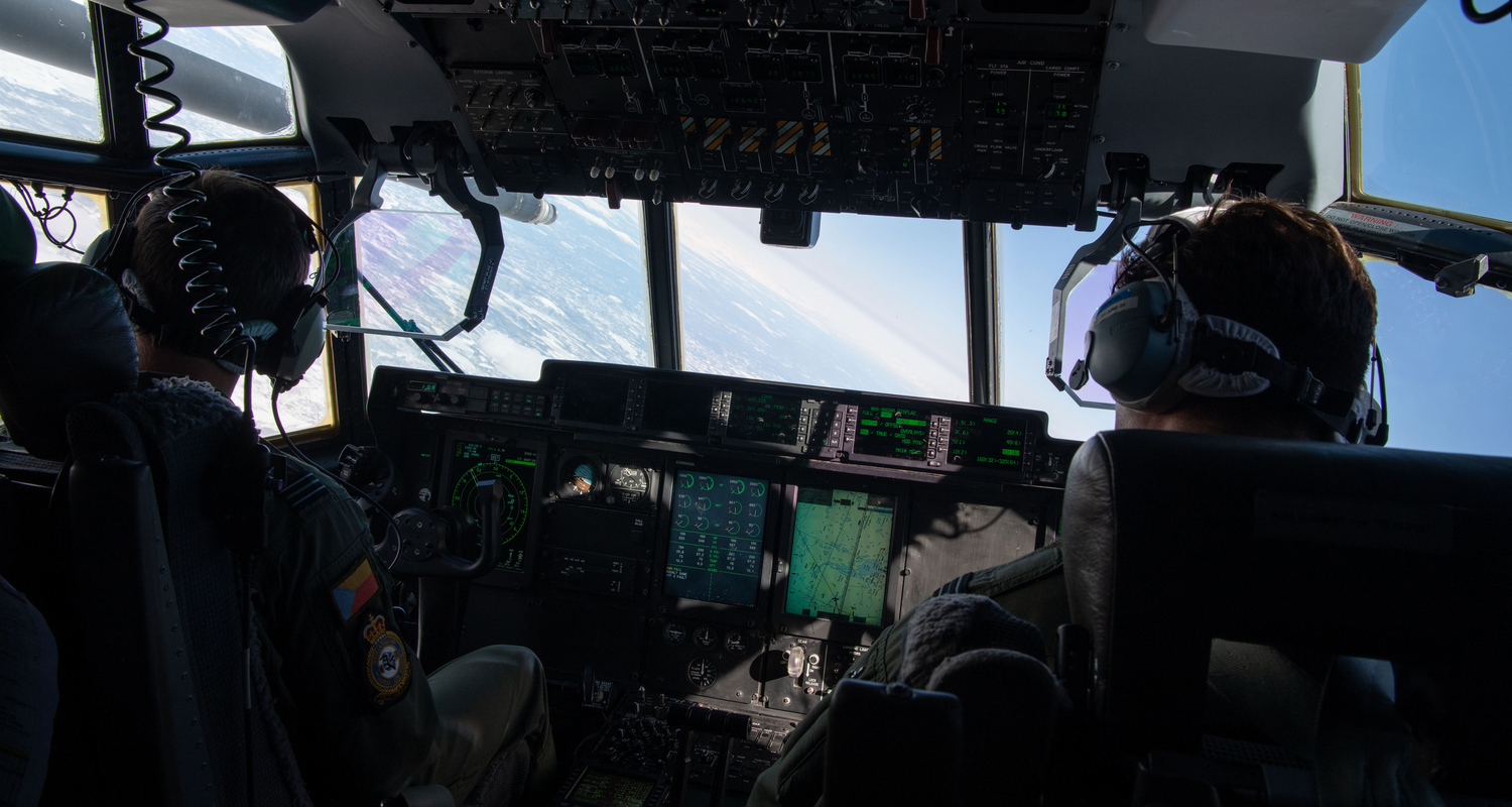 US Air Force pilots are flying an aircraft above the clouds. The photo was taken inside the cockpit, between the two uniformed personnel.