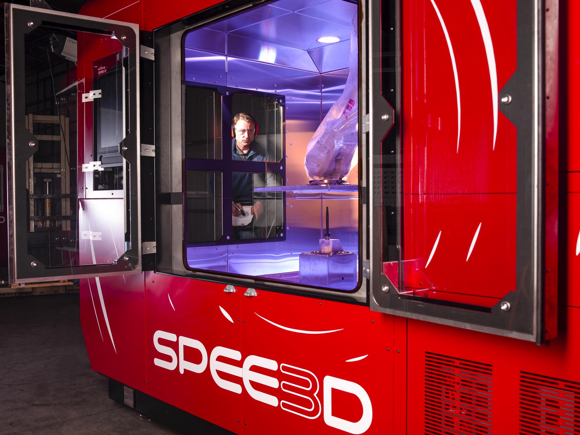 3D printing company SPEE3D's printing equipment, WarpSPEE3D, is seen on the photo. A man looks on in the background as he operates it, showing that the machine is the size of a small room, approximately 3 meters in height and 4 meters in length. Huge panels of glass between its sides show how it prints metal inside.
