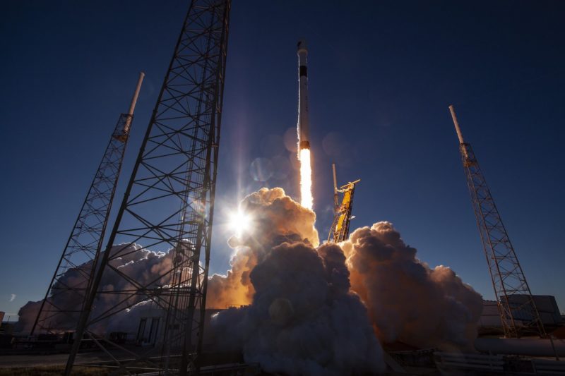 A rocket begins its launch. A huge cloud of smoke is seen below from it as it rises up, a red glow coming from its underside. The dark background is the night sky.