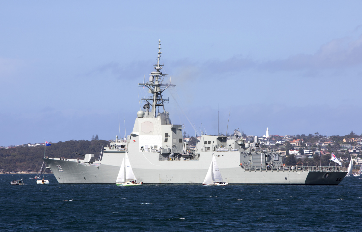 HMAS Hobart in Sydney Harbour prior to departing for her Mariner Skills Evaluation period. It has a white-grey hull with the number '39' painted on its left side in white. A coastal town serves as the background. The water is a deep blue.