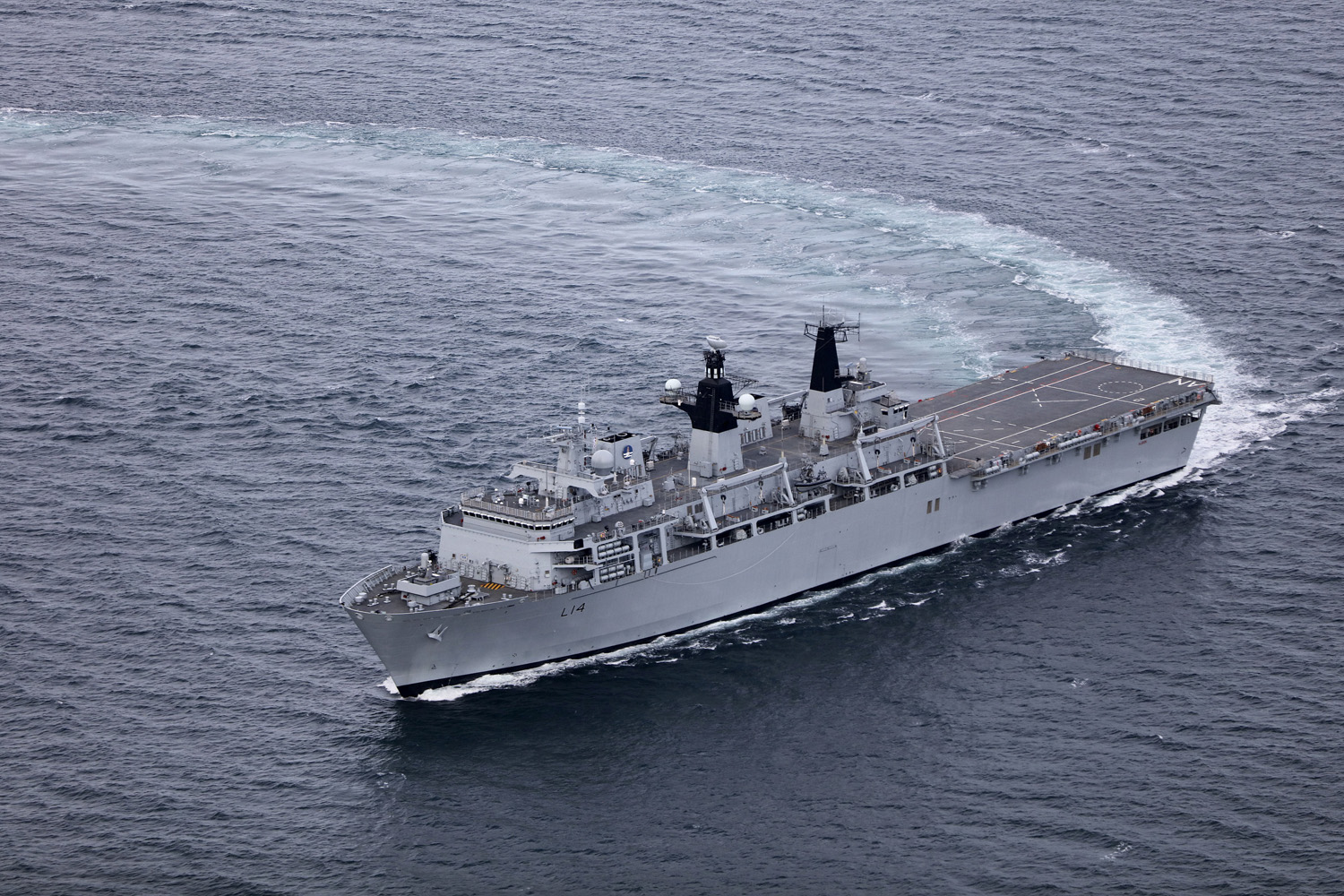 HMS Albion, an amphibious warfare ship, is seen sailing blue-grey waters. The photo appears to have been taken from above the waters, possible from an aircraft. The ship is painted grey and has 