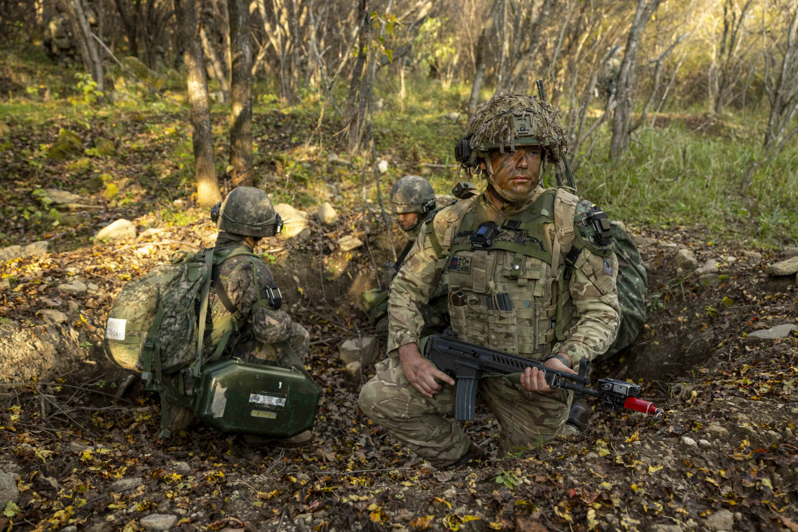 A soldier from C Company, the 1st Battalion Scots Guards checking their arcs alongside their Republic of Korean Army partners during the offensive phase during Exercise IMJIN WARRIOR. The soldiers are all wearing camouflage to blend in the mossy-green forest environment.