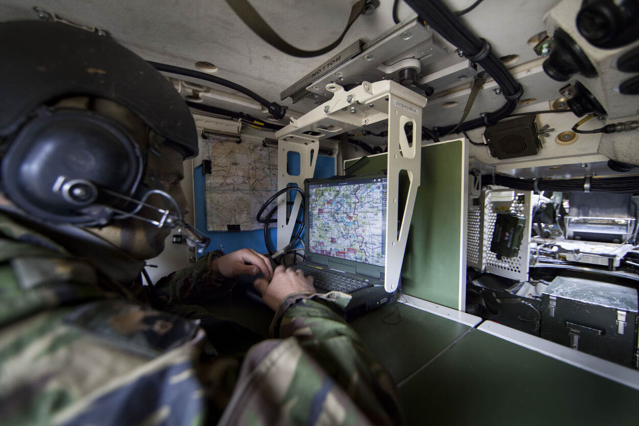 A military personnel, wearing camouflage and a microphoned headset, is seen using a laptop displaying a map. The photo appears to be taken inside a cramped military vehicle.