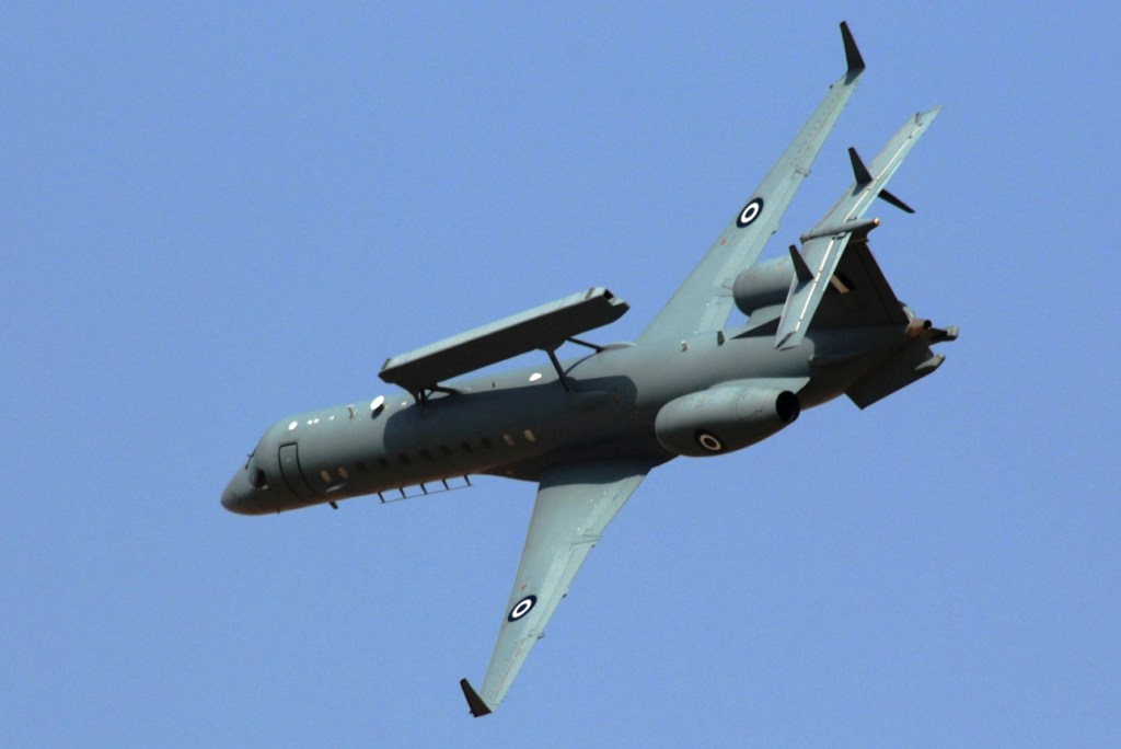 An Embraer 145 SA plane is seen flying. The background is a solid baby blue sky. The plane painted grayish green, with one bullseye pattern on each of the two wings and jet engines.
