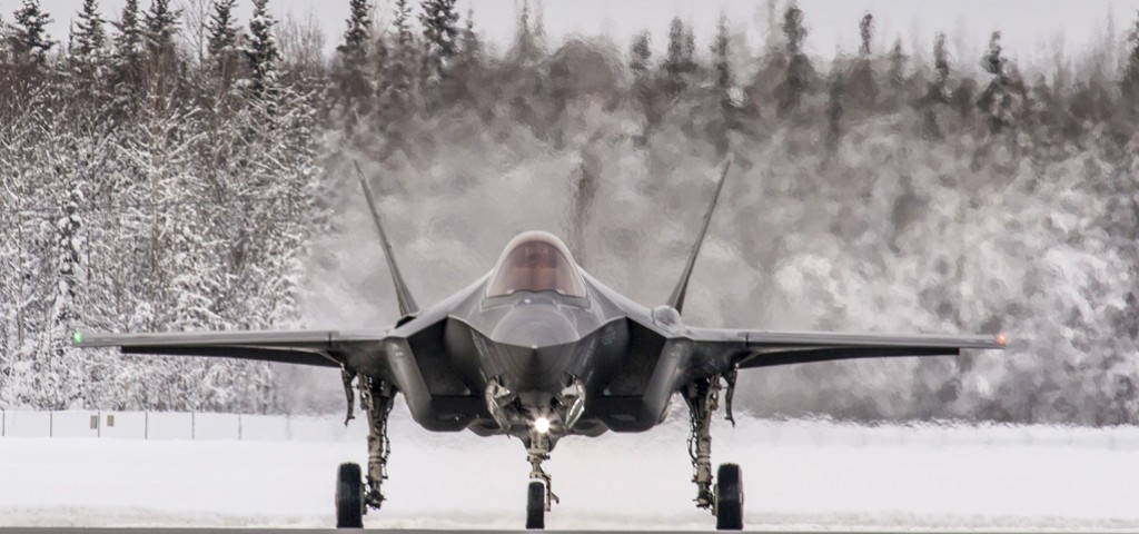 A black F-35 fighter jet plane is seen grounded, facing the viewer. The area it is in is snowy, with the ground completely covered in white snow. The background is a forest of snow-covered trees. The background on the back of the plane is blurred, presumably due to the heat distortion emitted by the plane.