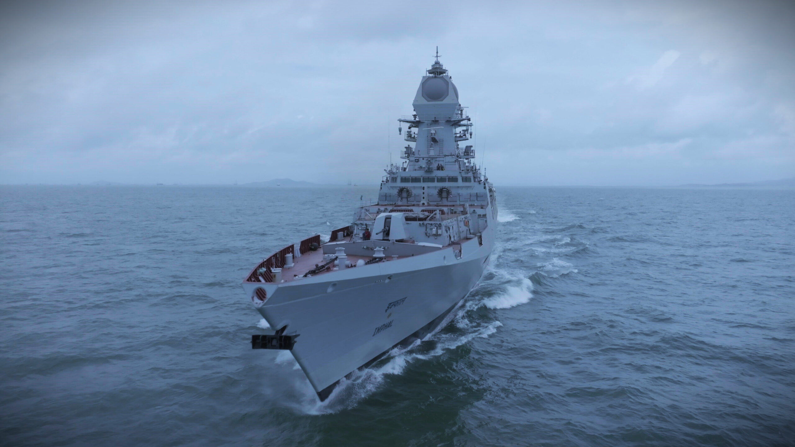INS Imphal, India's third Visakhapatnam-class stealth guided missile destroyer, is seen sailing in calm waters. The sky is dark and overcast, completely covered by heavy grayish-white clouds that give a gloomy feel to the whole image. The ship is painted gray.
