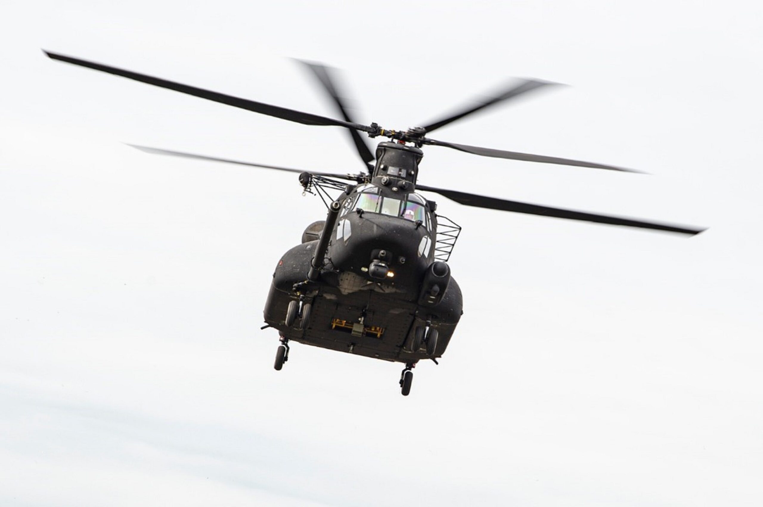 A black MH-47G Block II undergoing flight test is seen mid-flight. Its two overhead rotary blades are a little blurred as they spin. The background is a clear white sky.