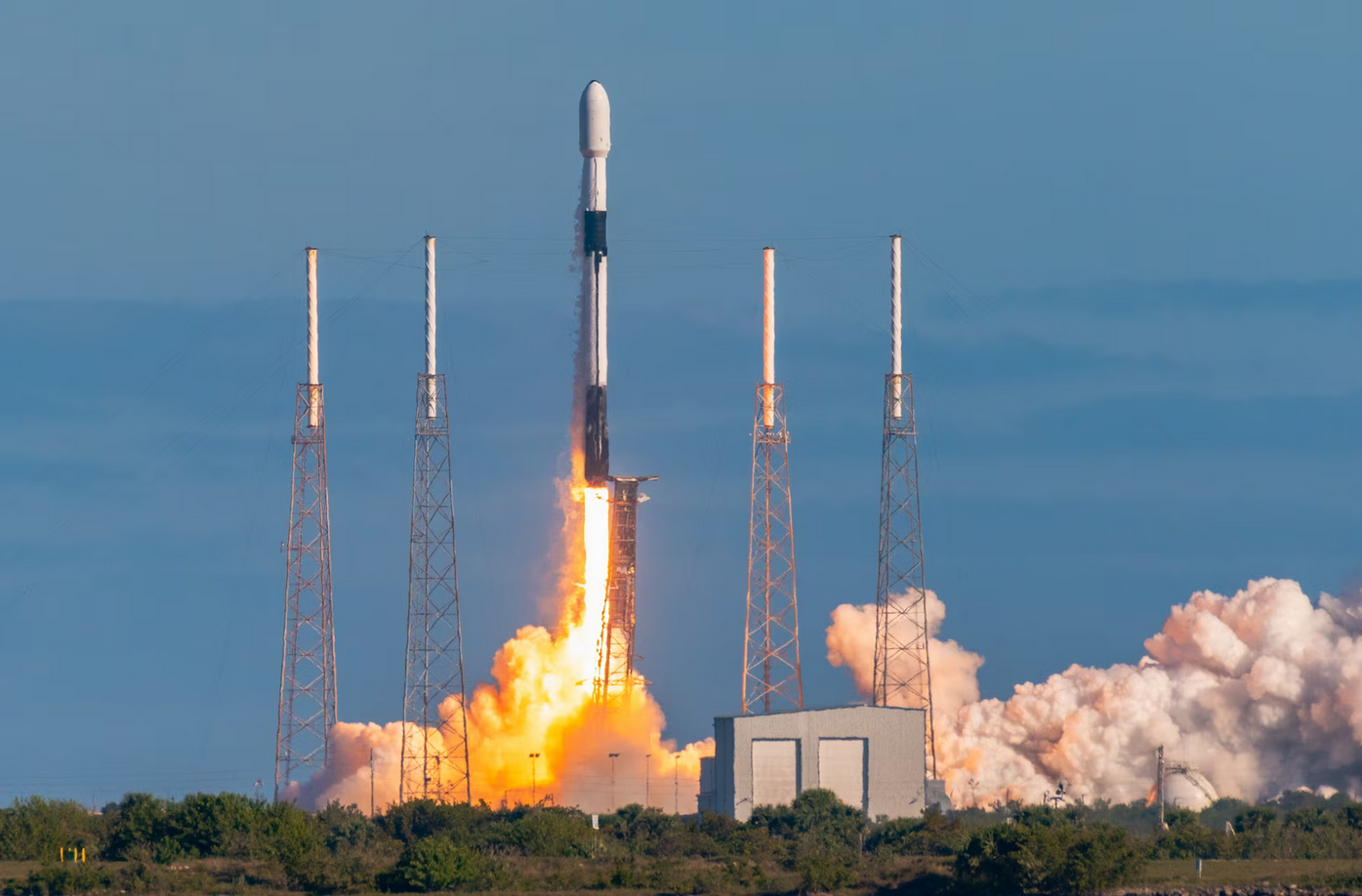 A Falcon 9 rocket launches from Cape Canaveral Space Force Station in Florida. Bright red-orange flames jut out from its back as it propels upward. Smoke is seen covering the lower right side of the image. The background is a calm blue sky.