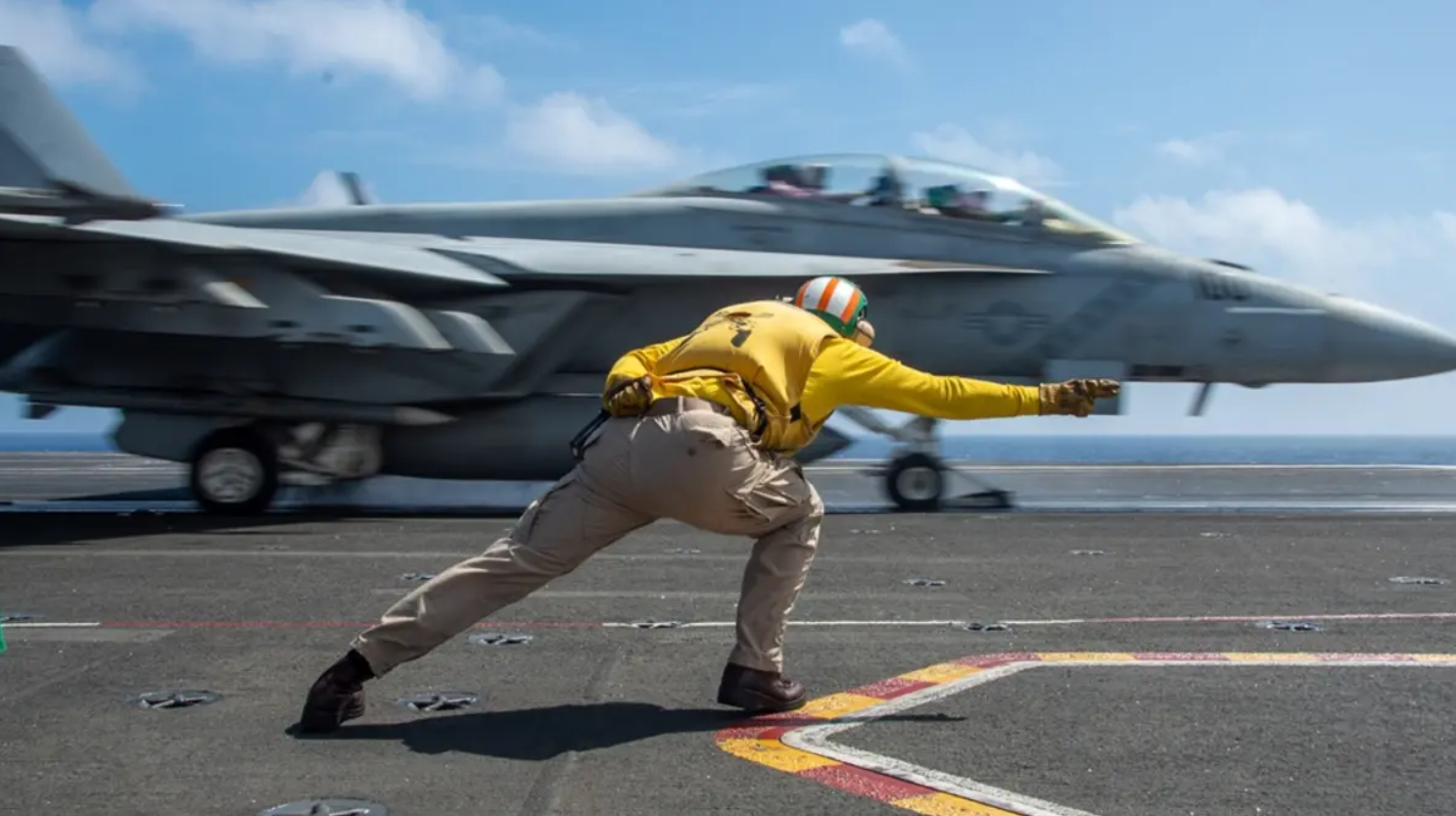 A military personnel is seen guiding a jet plane in what appears to be an on-sea landing system. The personnel is wearing a yellow jacket and brown khakis. The fighter plane is colored gray. The background is a cloudy blue sky, with the ocean water below.
