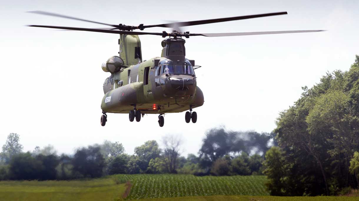 An medium-to-heavy lift CH-147F Chinook helicopter is seen hovering over a green landscape with trees sprawling in the background and the far right side of the image. The helicopter is colored a dark green and grey camouflage. Its front and back propellers are due to its fast rotations.The far background is a bright white sky.