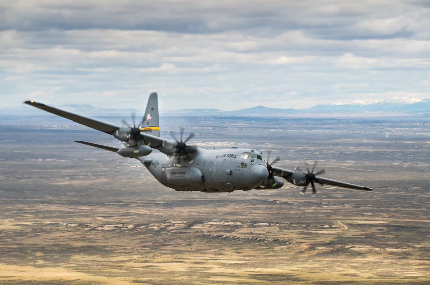 A C-130 Hercules flies over a sandy plains. The plane is colored gray, with a yellow stripe on its tail. It has 