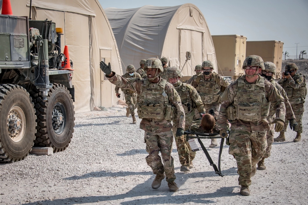 Four camouflaged US Soldiers at the Al Udeid Air Base in Qatar are seen carrying a soldier on a stretcher in what appears to be a military drill. On the back, at least six more soldiers are seen tailing them. On the background is two brown tents and two brown military trucks. On the left, the back of another truck is seen.