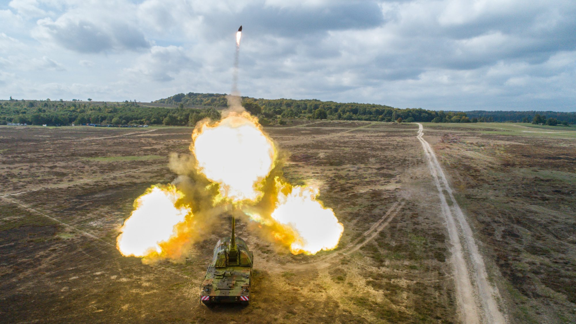 A Panzerhaubitze 2000 tank is seen firing a 155mm ammunition at the Ravlunda test field in Sweden. Three balls of blazing fire are seen jutting out of the tank's gun barrel, with the ammunition flying off upwards. The test field is a greenish-brown plains dotted with small plant growths. In the distant background, a hilly landscape covers the horizon. The blue sky is full of light-gray clouds.