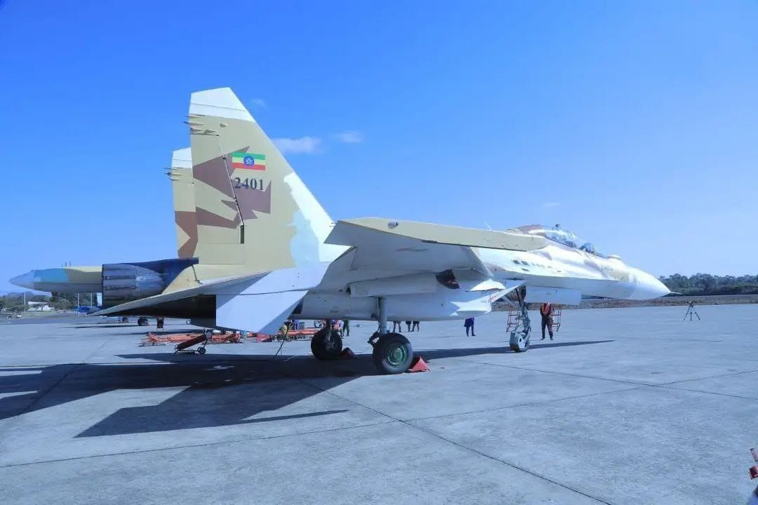 Two Sukhoi Su-30 planes are seen stationed on what appears to be a military air base. The planes are painted white, except for its camouflaged tails. The plane in front has 