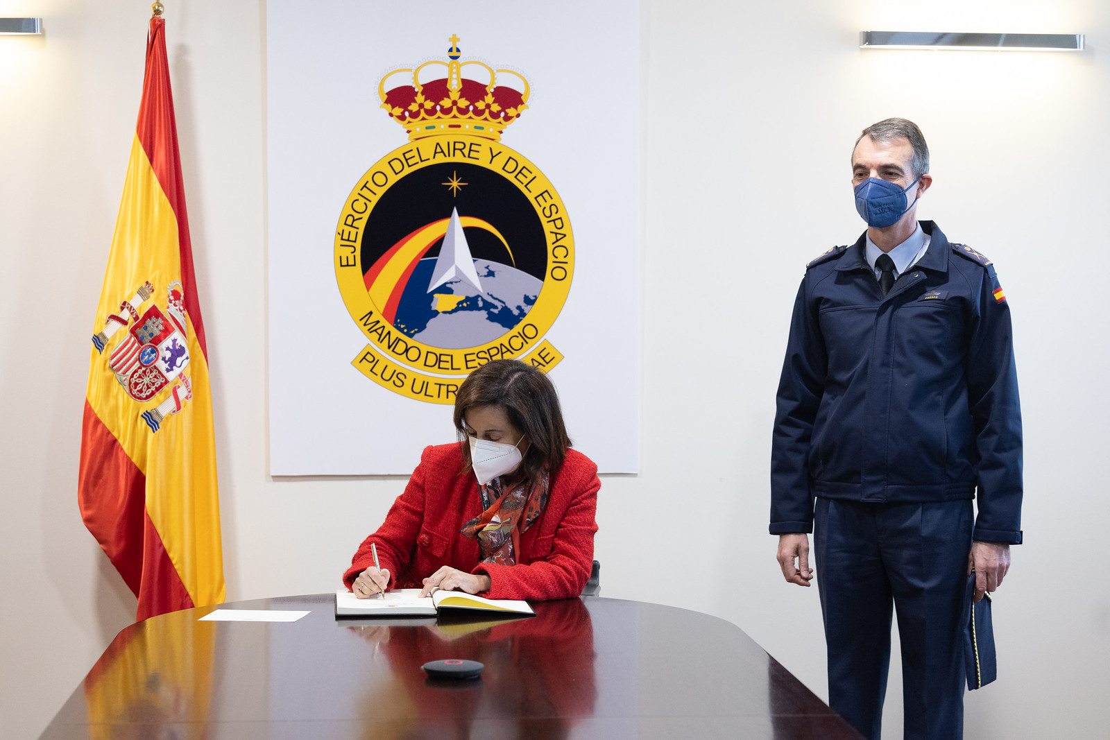 Span's Defense Minister Margarita Robles is seen signing a document inside what appears to be a conference room. An office personnel is seen standing by her left. The Spanish Air and Space Force emblem is displayed on the background, and the Spanish flag is positioned to her right.