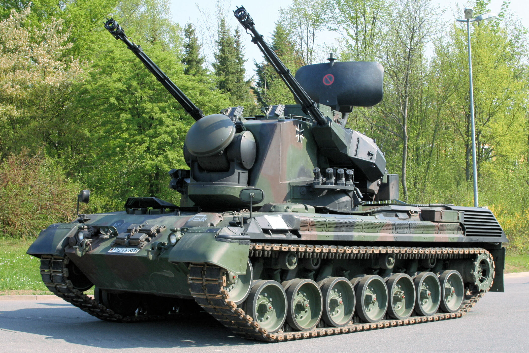 A Gepard anti-aircraft gun system is seen parked on the side of an asphalt road. The tank is painted with a dark green and brown camouflage. Its two gun barrels are pointed to the upper-left, angled at around 45 degrees. The background is a landscape of green-leaved trees.