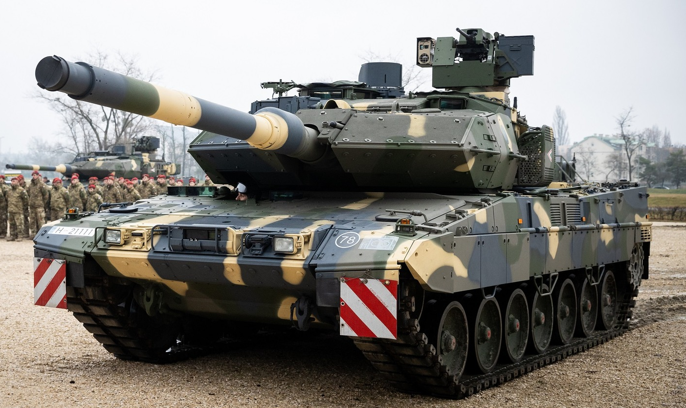 A camouflage-painted Leopard 2A7HU tank is seen stationed in a sandy area. Another tank is parked on the far left side of the background, with camouflage-wearing soldiers standing in attention in front of it. Some trees are seen poking out the far back of the image. The sky is a dull, foggy white.