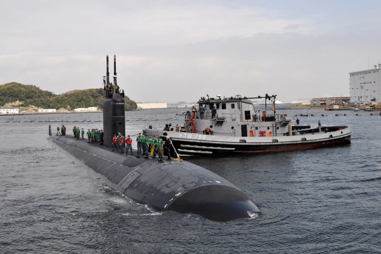 The USS Columbus submarine is seen partially submerged in the waters of what appears to be a pier. A boat with 
