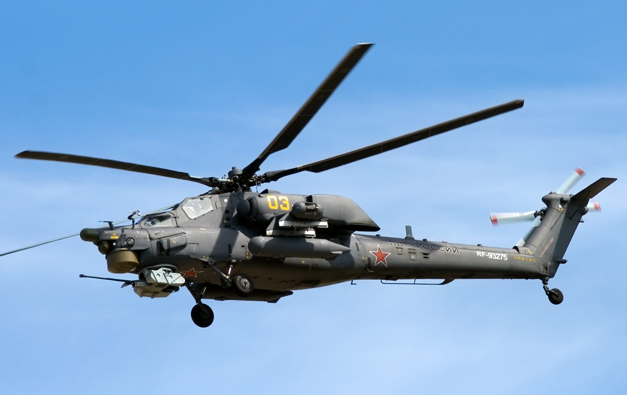 A Mil Mi-28 helicopter is seen hovering on the clear blue sky. On its gray-green hull, the number 