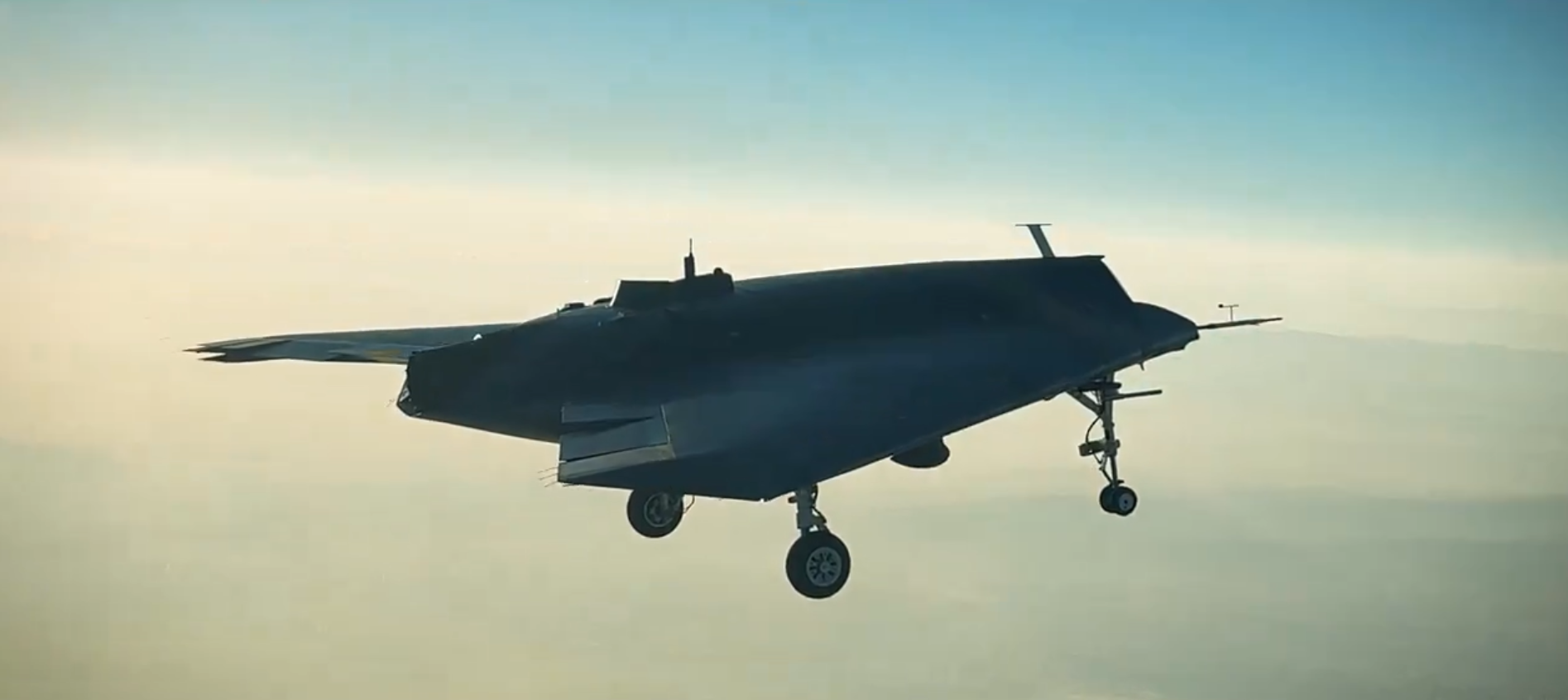 A gray Turkish Aerospace Industries Anka-3 unmanned combat aerial vehicle is seen flying above the clouds. The warplane is largely flat-shaped, with its four landing wheels jutting out underneath. The background is a light blue sky, with white clouds reflecting the sun's orange rays below.