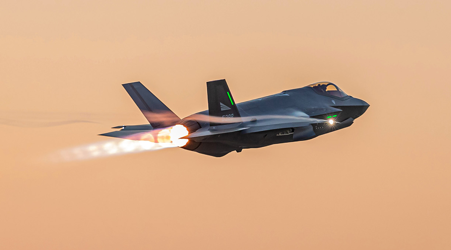 A black Royal Norwegian Air Force F-35 plane is seen flying in an orange, sunset-tinged sky. The plane's wing has 
