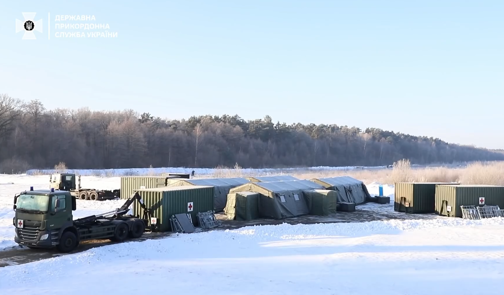 Hospitainer's Role II Hospital, a mobile, inflatable building, is seen set up in a snowy field outside a forested area in the background. Two container trucks are parked outside of it, and two cargo truck containers are also positioned on the far right of the field.