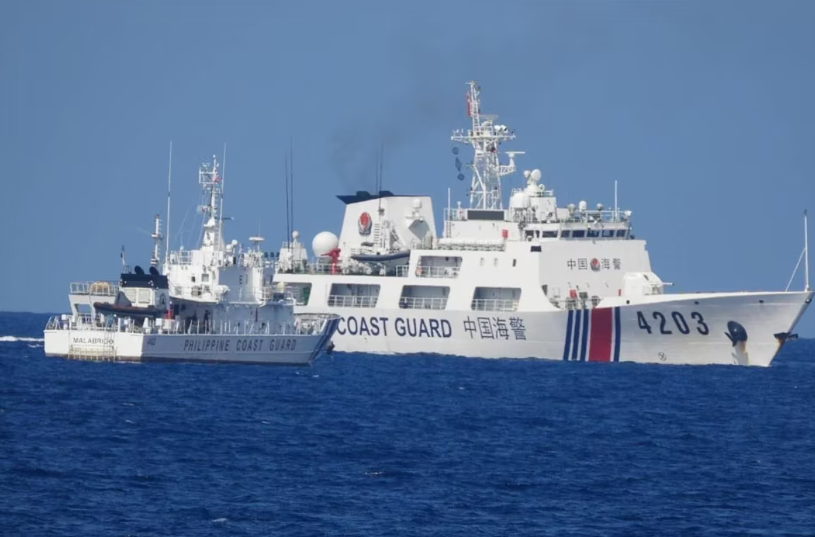 A white Philippine Coast Guard ship is stationed in the waters of the West Philippine Sea, facing a larger Chinese Coast Guard ship. The sea is a deep blue color, and the sky is a dull blue.