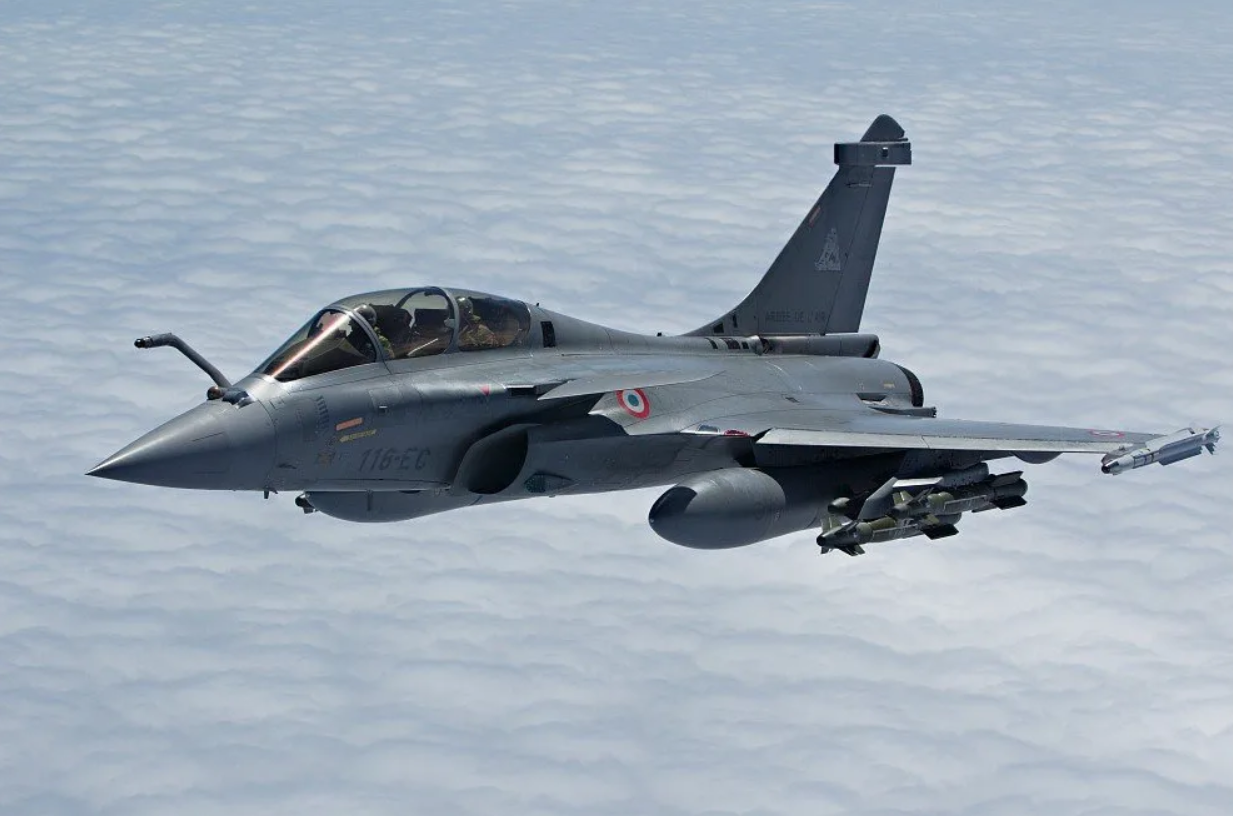 A gray Dassault Rafale fighter jet is seen flying above a sea of white clouds. It's equipped with what appears to be two missiles under the middle of its left wing, and a smaller one on its tip.