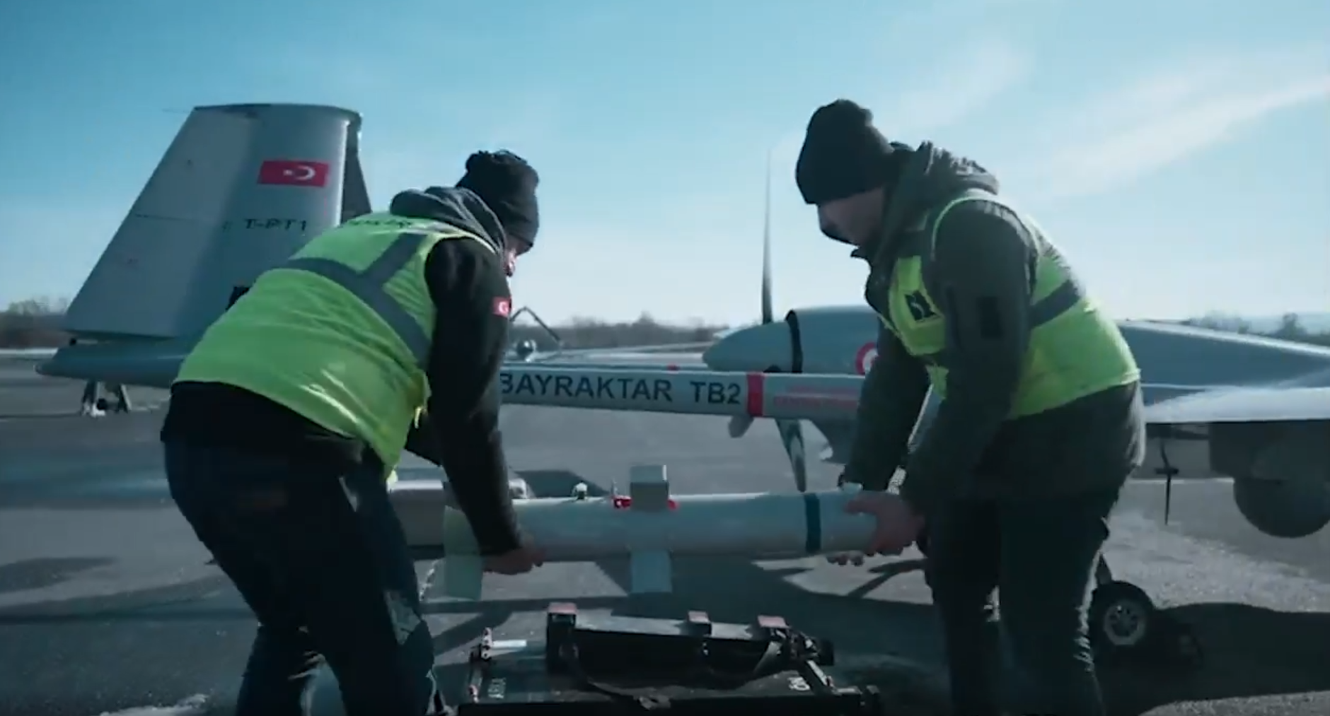 Two individuals are carrying a Desert Sting 16 bomb, preparing to install it under the wing of a Bayraktar TB2 drone. The individuals are wearing neon-green high-visibility vests. Both the bomb and the drone are painted gray. They appear to be set outside a hangar. The background is a clear blue sky with some thin white clouds.