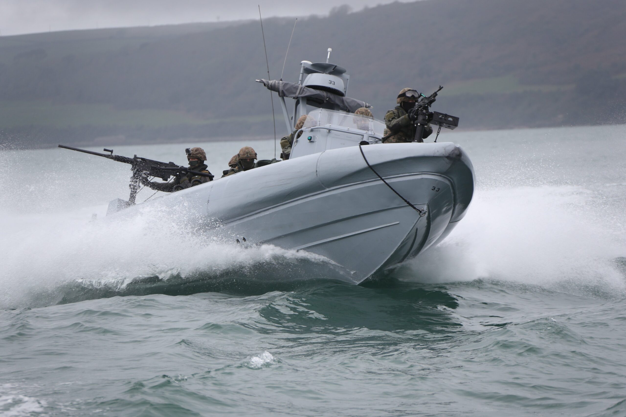 The Commando Raiding Craft is seen in action in Plymouth Sound. It's seen moving in a coastal body of water, with at least six personnel on the craft. Two soldiers are manning what appears to be machine guns, one in front of the boat and the other in the back. In the background, a coastal land mass is seen amid foggy weather.