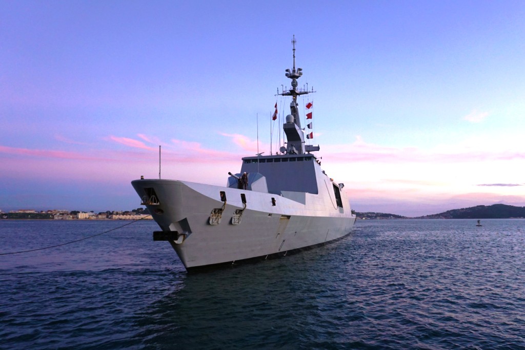 The French frigate Aconit (F713) is seen docked in what appears to be a pier. Its hull is painted gray. A rope is seen tied to its front. It has flags of varying colors decorated beside its mast. The water is a deep, dark blue. The background is a blue sky with purple clouds, reflecting the sun out of frame.