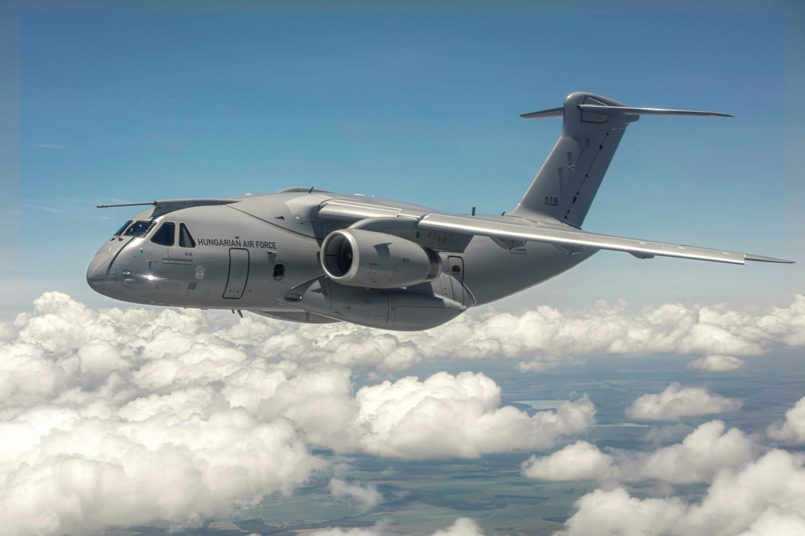 An Embraer C-390 Millennium aircraft is seen flying above the clouds. The gray aircraft has the words 