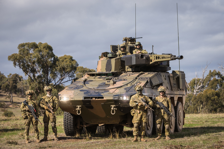 Rheinmetall Boxer Combat Reconnaissance Vehicle is seen with its headlights turned on in a grassy field. The armored 8x8 vehicle is painted in camouflage. Two pairs of camouflaged soldiers holding rifles flank its sides. On top of the vehicle, two more soldiers are seen. The sky is gray and filled with dark clouds, and some trees are seen scattered on the background.