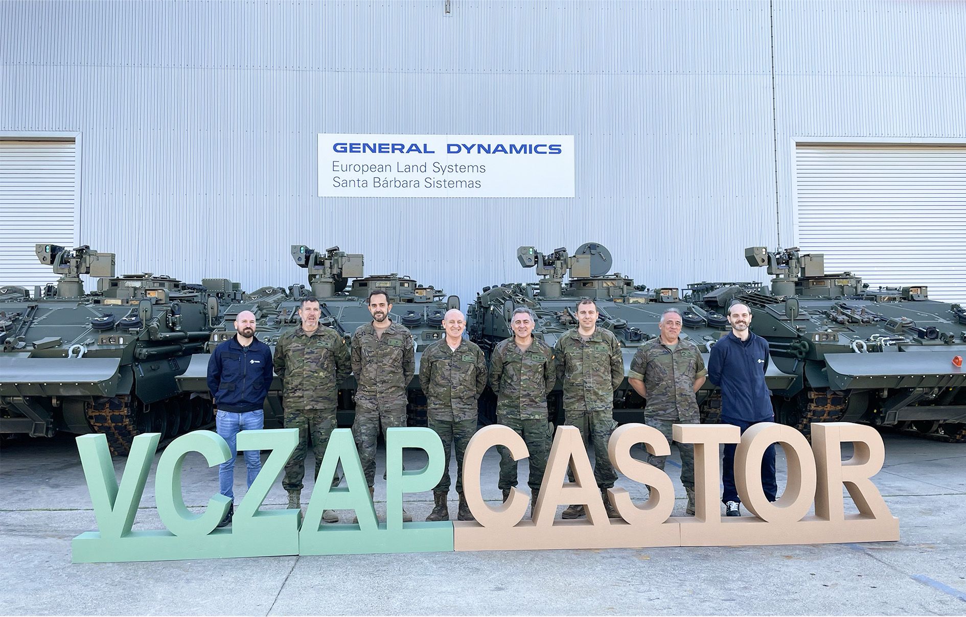 Six uniformed Spanish Army personnel stand in front of four ASCOD Castor armored combat vehicles. They are joined by two civilians, likely from General Dynamics. In front of them are letter standees that spell out 