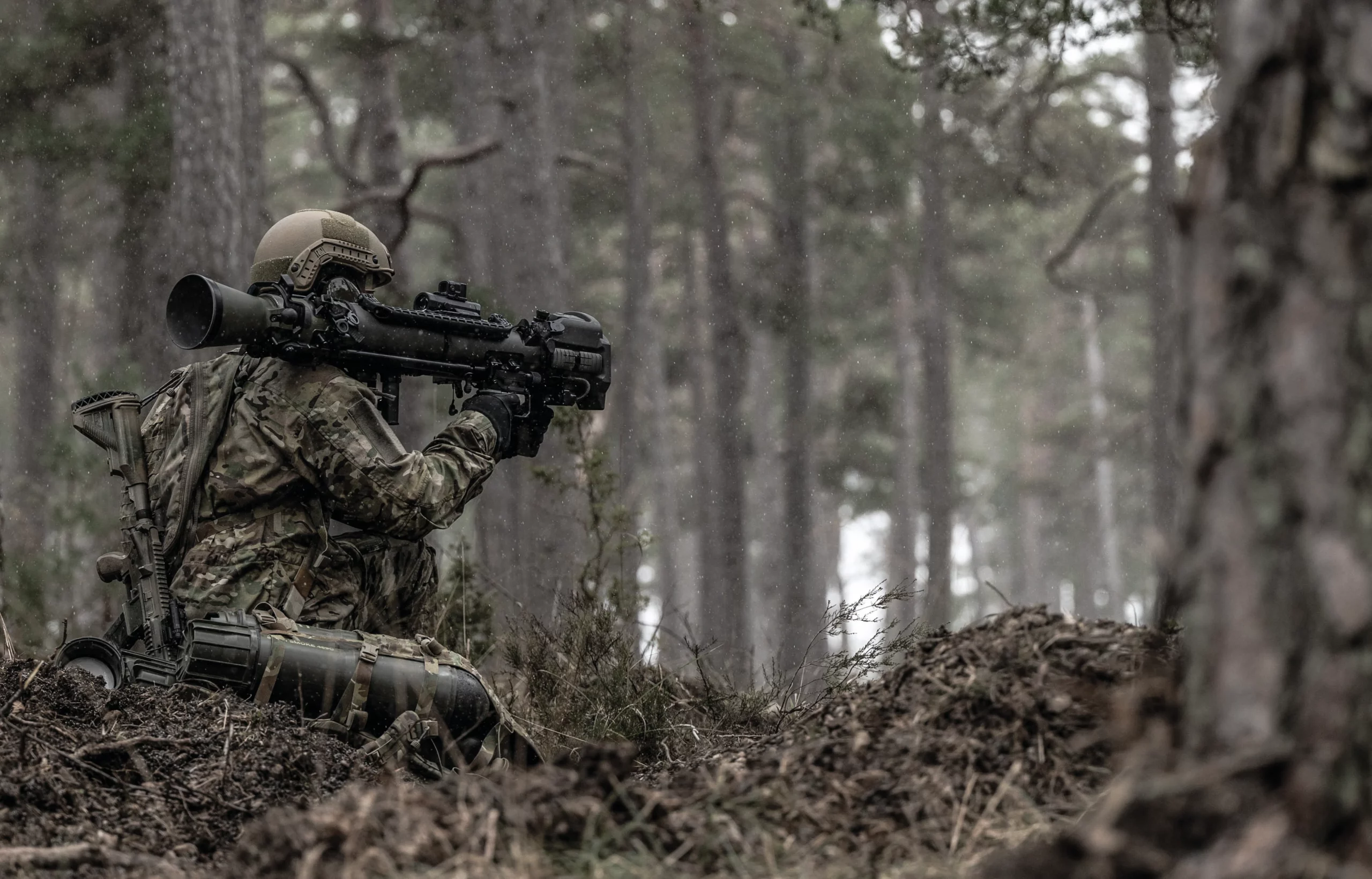 A soldier is seen with his back turned, seemingly laying low in a brown, forested area. He is wearing camouflage. On his right shoulder, a black Carl-Gustaf rifle rests as he props it up to point forward.