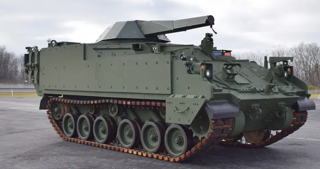 The Armored Multi-Purpose Vehicle Mortar Carrier prototype is seen in a large parking area. The tracked armored vehicle has a turret jutting out from its body. The entire vehicle is painted dark green.