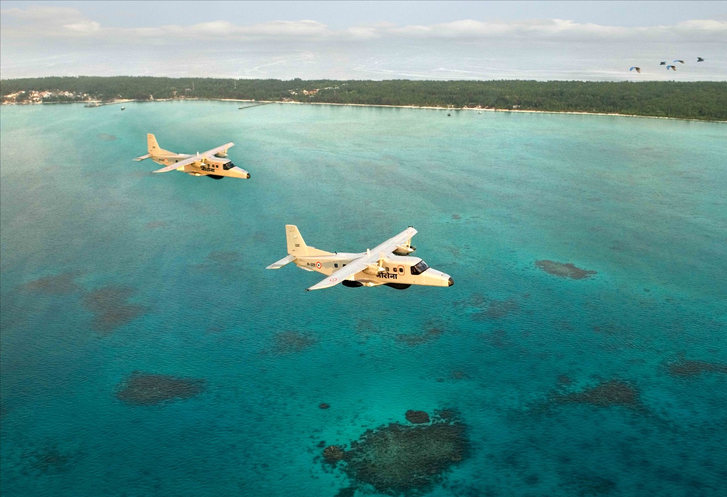 Two of the Indian Navy's Dornier 228 aircraft are seen flying above calm light blue-green waters. The planes are painteed white, with phrases painted in black in Devanagari (Indian script).. In the far background, a shorescape full of trees are visible, with some birds flying on the far upper right corner of the image.