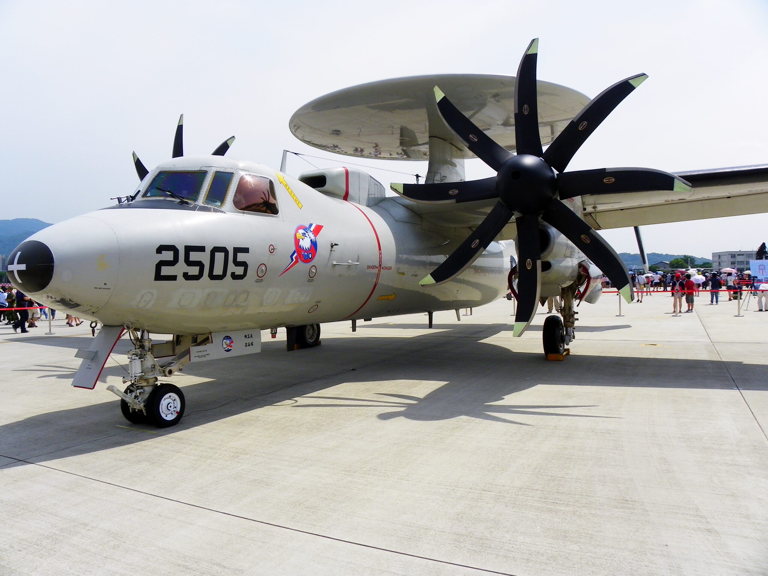 A Northrop Grumman E-2K Hawkeye plane appears to be on display at a wide-open area similar to a runway. It is cordoned off by a red-velvet stanchions seen in the back. The gray plane has 