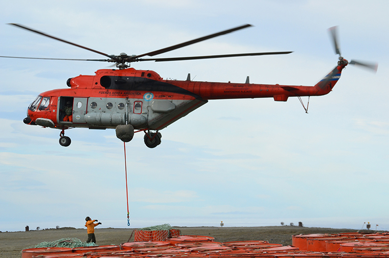 Argentina's Mi-171E helicopter, H-95, is seen flying above a green plains. An orange cable with what appears to be a hook contraption on its end dangles from its body. The helicopter is painted orange on its upper half and outside the cockpit, and gray on its lower half. Its body has many writings and decals on it, including 