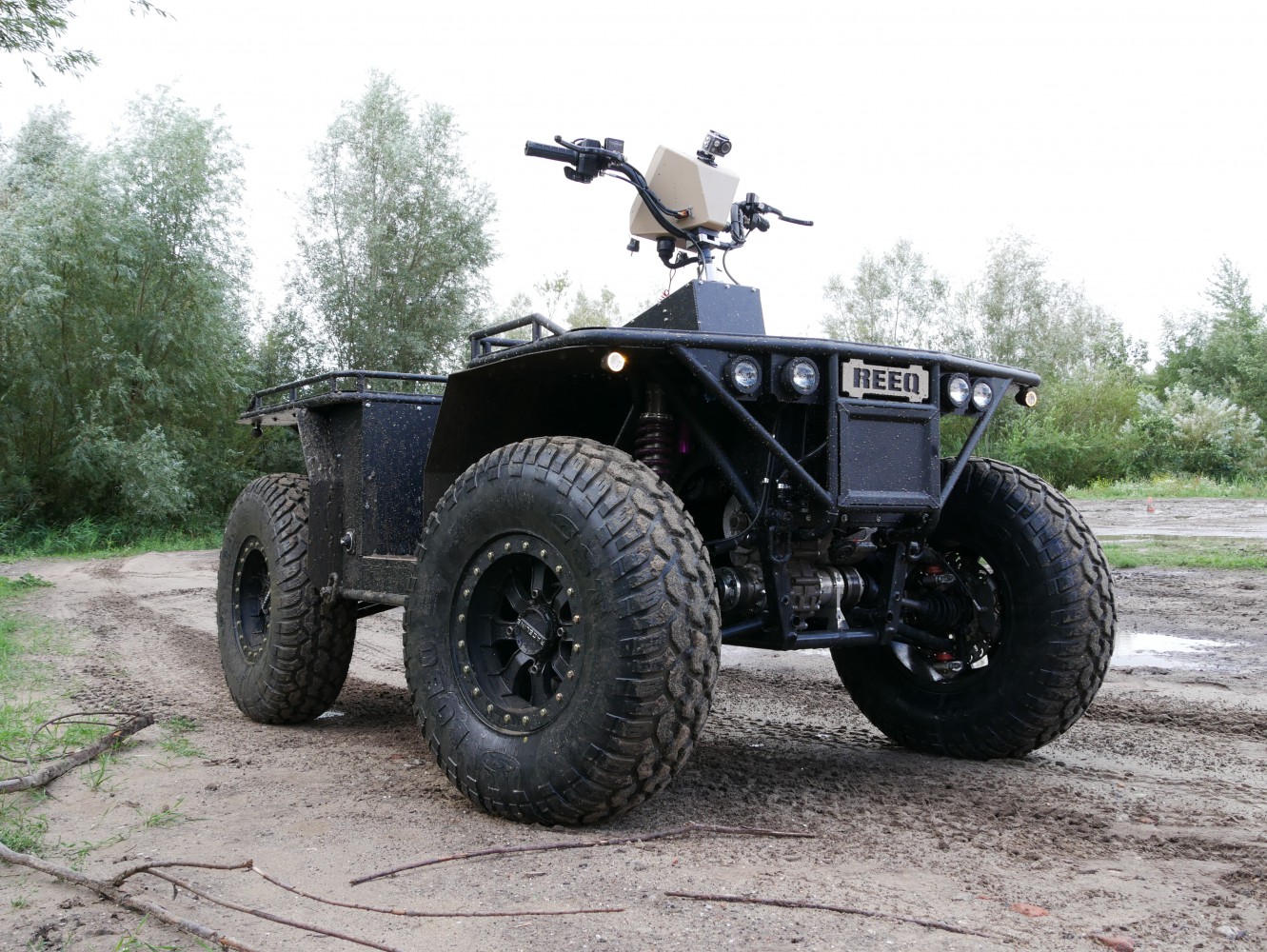 A REEQ Rex Quad is seen parked in a muddy field with trees and shrubbery in the background. The vehicle is a 4-wheel off-road vehicle with large wheels and a minimalistic chassis. Most of its parts are painted black, except for the logo in front and the casing of the screen in the middle of the handlebars, which are both dirty brown.