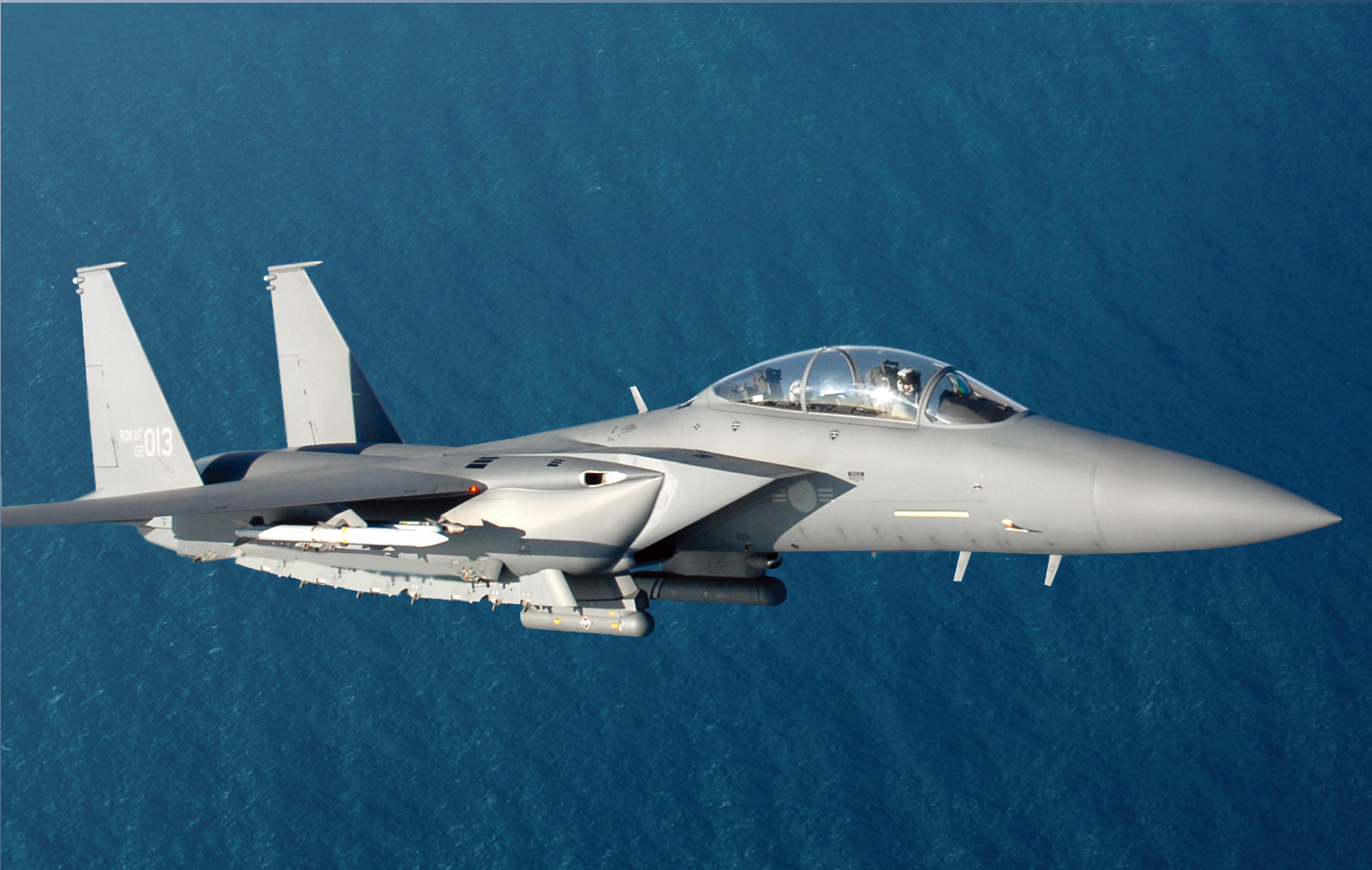A Republic of Korea Air Force F-15K fighter plane is seen flying above a body of water. The fighter plane is painted gray and has a sleek body to make it more aerodynamic. The number 