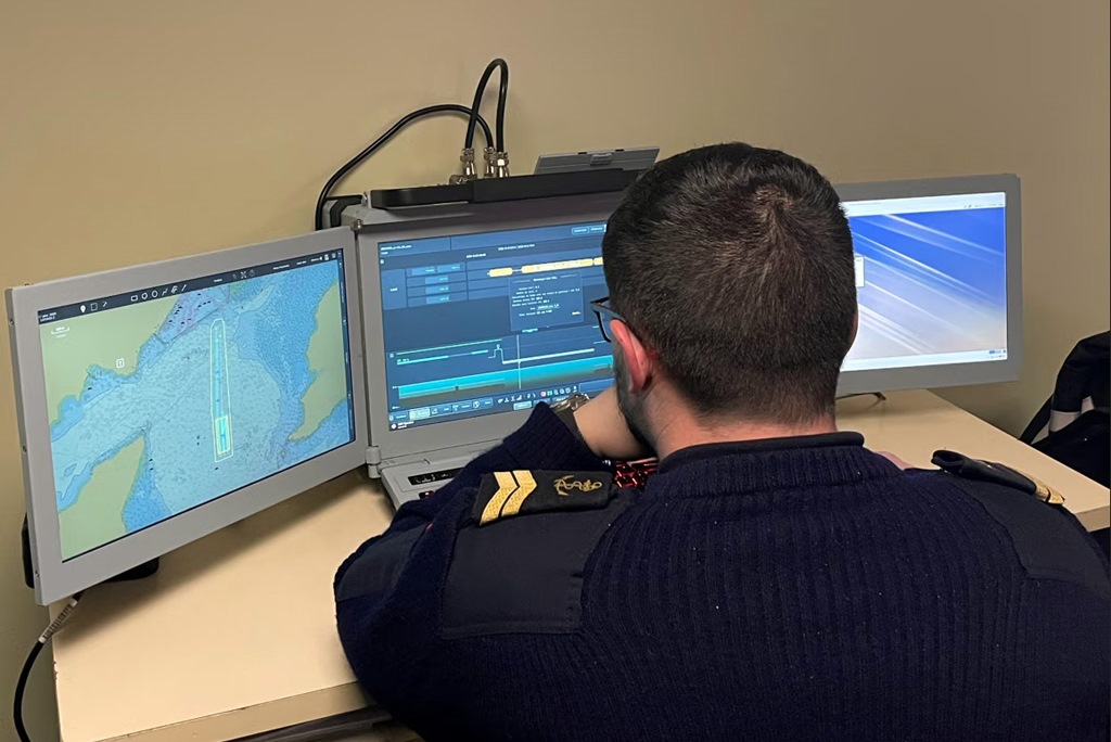 A uniformed French Navy personnel has his back turned against the camera as he sits in front of a computer system with three computer screens propped up on a table. The screens show what appears to be the interface of Thales' Expeditionary Portable Operations Centre (e-POC). The left screen displays a topographic map, and the middle screen shows various data through lines and graphs. The screen on the right appears to be displaying a blue-violet wallpaper.