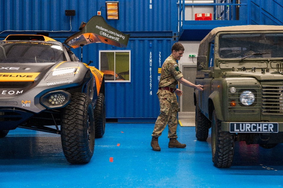 A British Army land rover with an electric engine is seen parked on the right side of the image, right beside a NEOM McLaren Extreme E racecar on the left. A uniformed army personnel is seen reaching for the land rover's door, as if opening it. The rover is painted brown-green, with a plate in its front that reads 