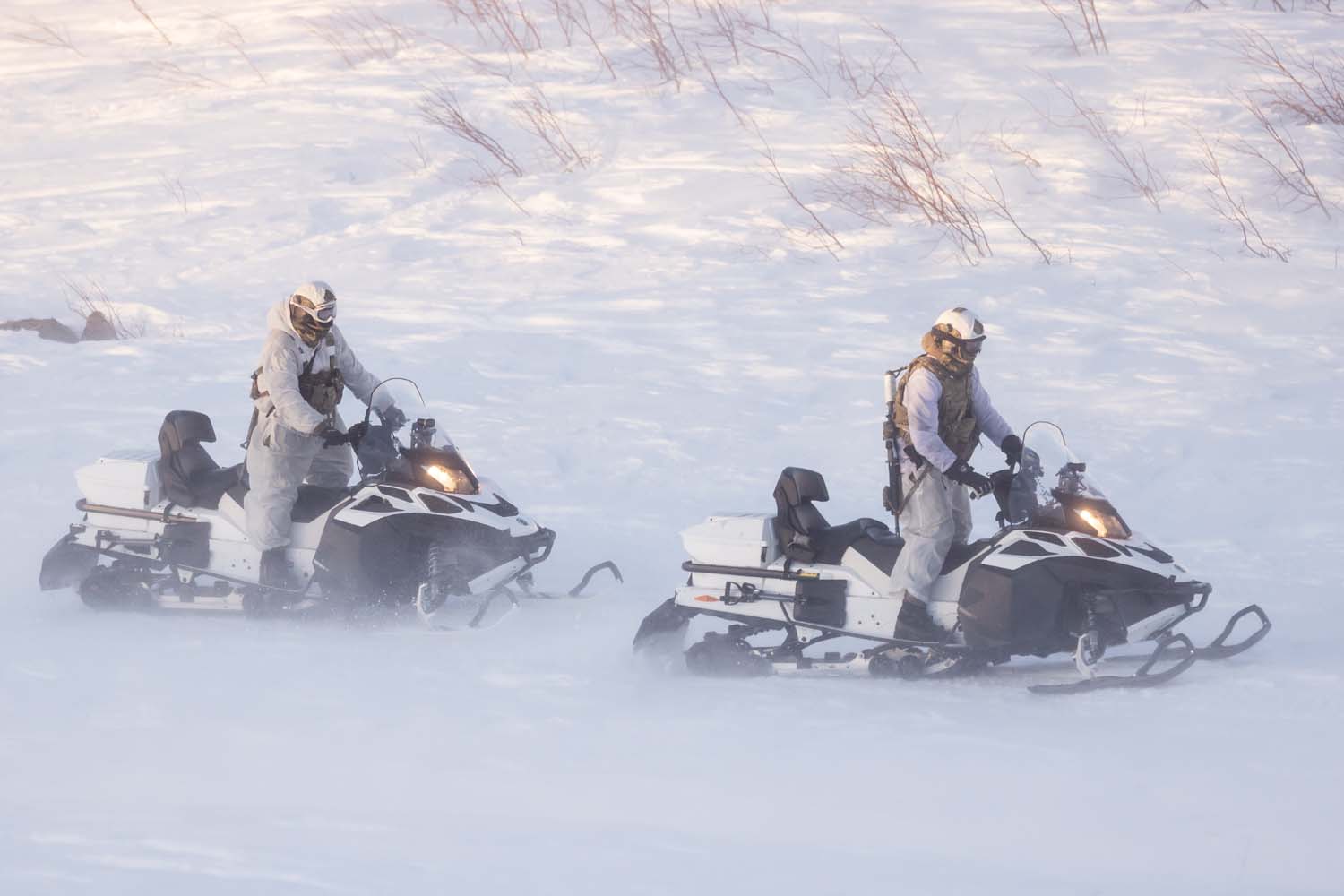 Two soldiers are seen operating two black-and-white Lynx Brutal Over Snow Reconnaissance Vehicles in the middle of a snowfield. The soldiers are wearing white as camouflage to blend in well with their surroundings.