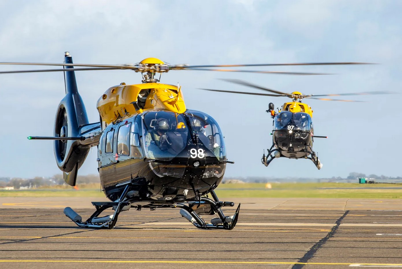 Two Airbus H145 helicopters are seen operating in what appears to be an aircraft runway. The helicopters are painted black, with its rotary blades components and base painted yellow. The one in the foreground has the number "98" painted in white in front of its cockpit, while the one in the back has "97".