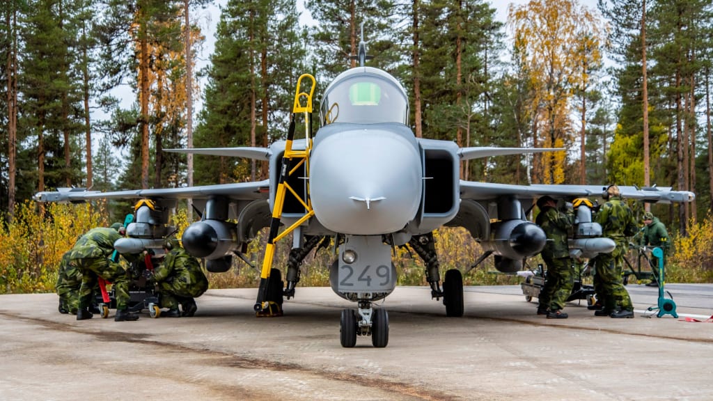 A Gripen C-series plane is seen being serviced by uniformed personnel while parked in the ground. All seven personnel are wearing green camouflage uniforms. The plane is painted gray with the number 