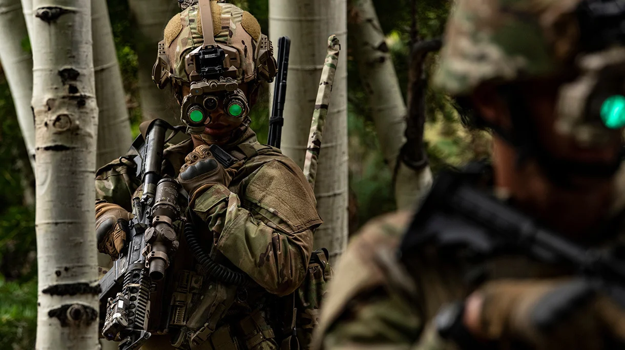 Two soldiers are seen wearing Enhanced Night Vision Goggle-Binoculars (ENVG-Bs). One soldier is on the far right foreground, out of focus. The other one is on the left side of the image. Both are wearing camouflage to match the wooded environment. The binoculars are strapped to their helmets, and its eyepiece are glowing bright green.